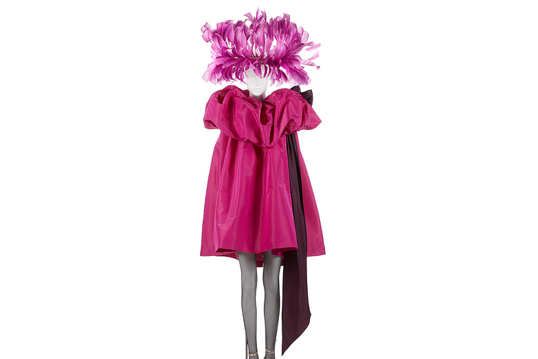Mannequin wearing a knee-length voluminous, bright pink dress with a large ruffle around the shoulders and a feathered headpiece.