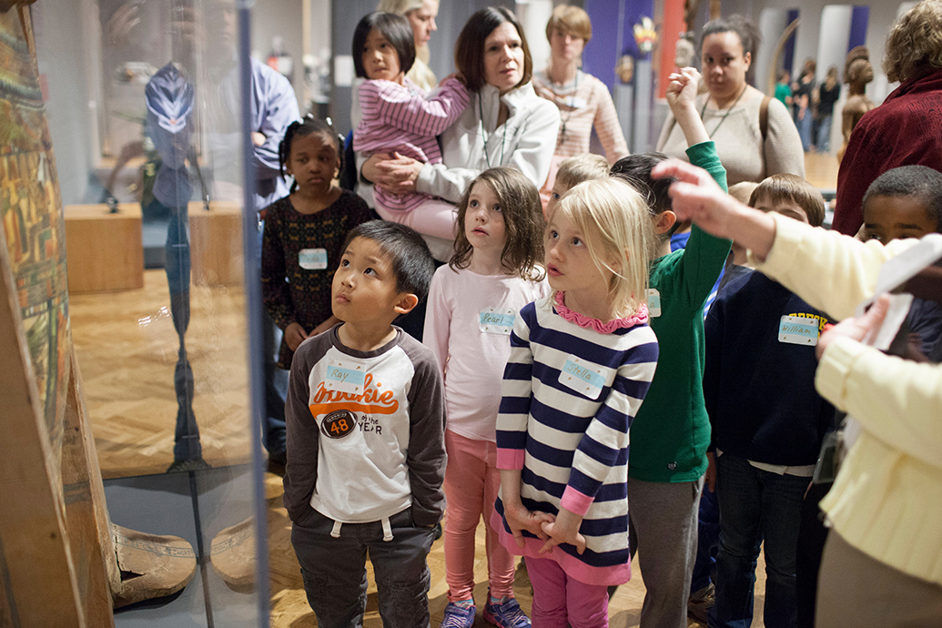 A group of kids listening to a speaker, and looking at an object in vitrine.