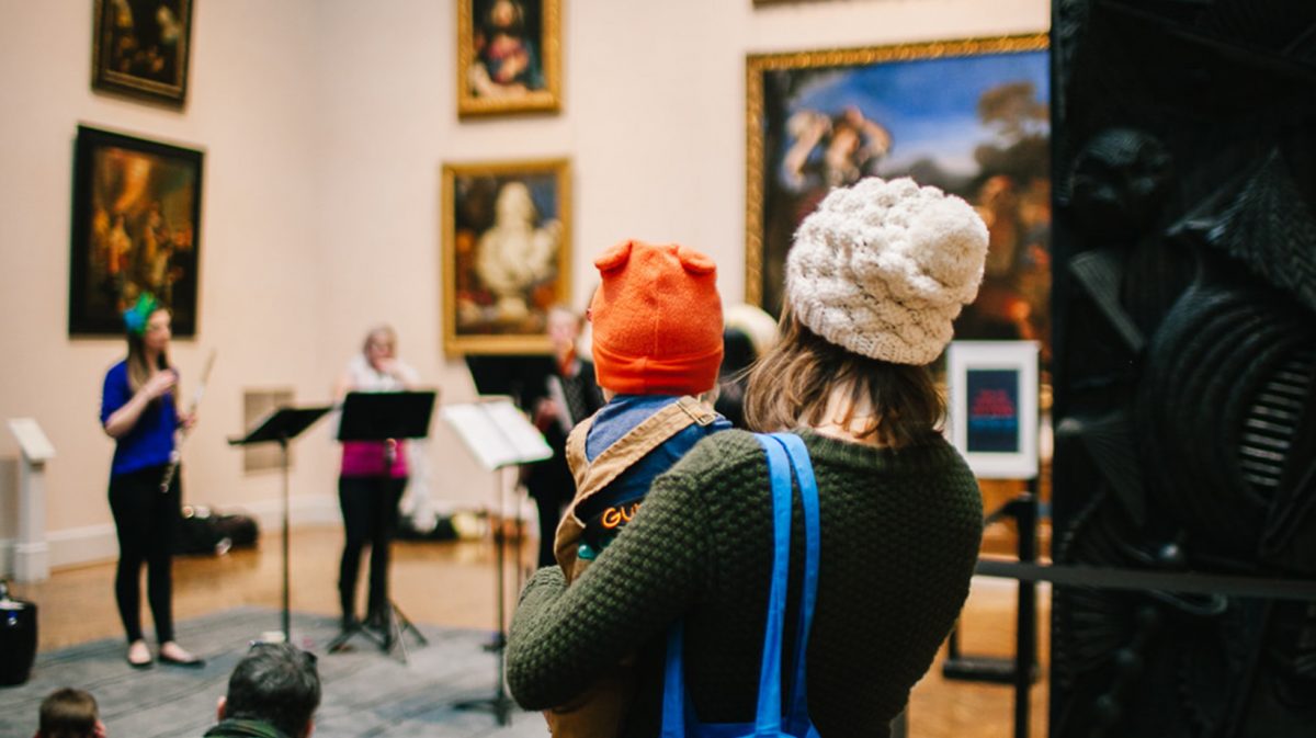 Woman holds young child while watching a gallery performance.