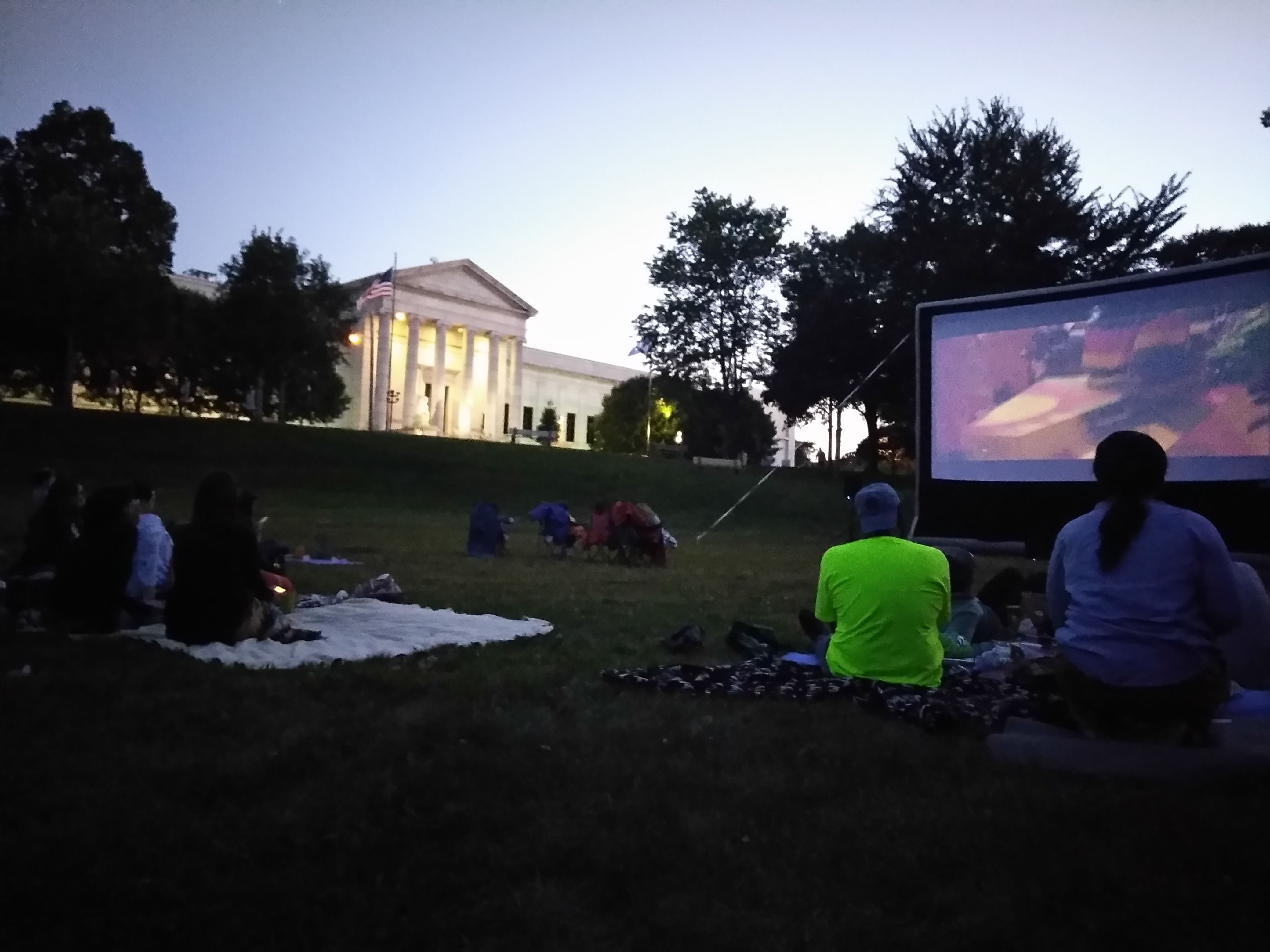 A large movie screen and small groups of people watching in the park across the street from Mia.