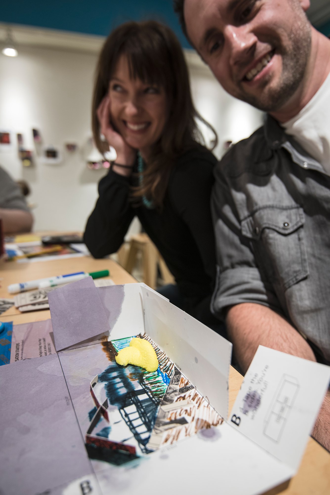 Two adults in grey shirts smile with their hand made collage artwork in Mias studio classroom