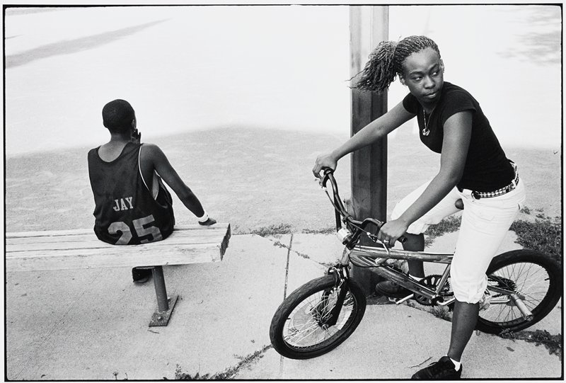 A black and white photograph of two kids - one is on a bike looking to their left and the other has their back to the camera.