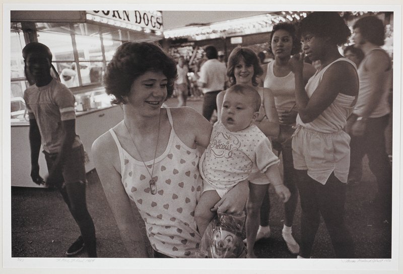 A woman holding a baby in the center of a crowd of people near a corn dog stand