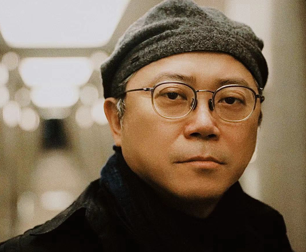 This is a close up photo of art director, Tim Yip. He is wearing glasses, in an all black outfit with a gray tweed flat caps.