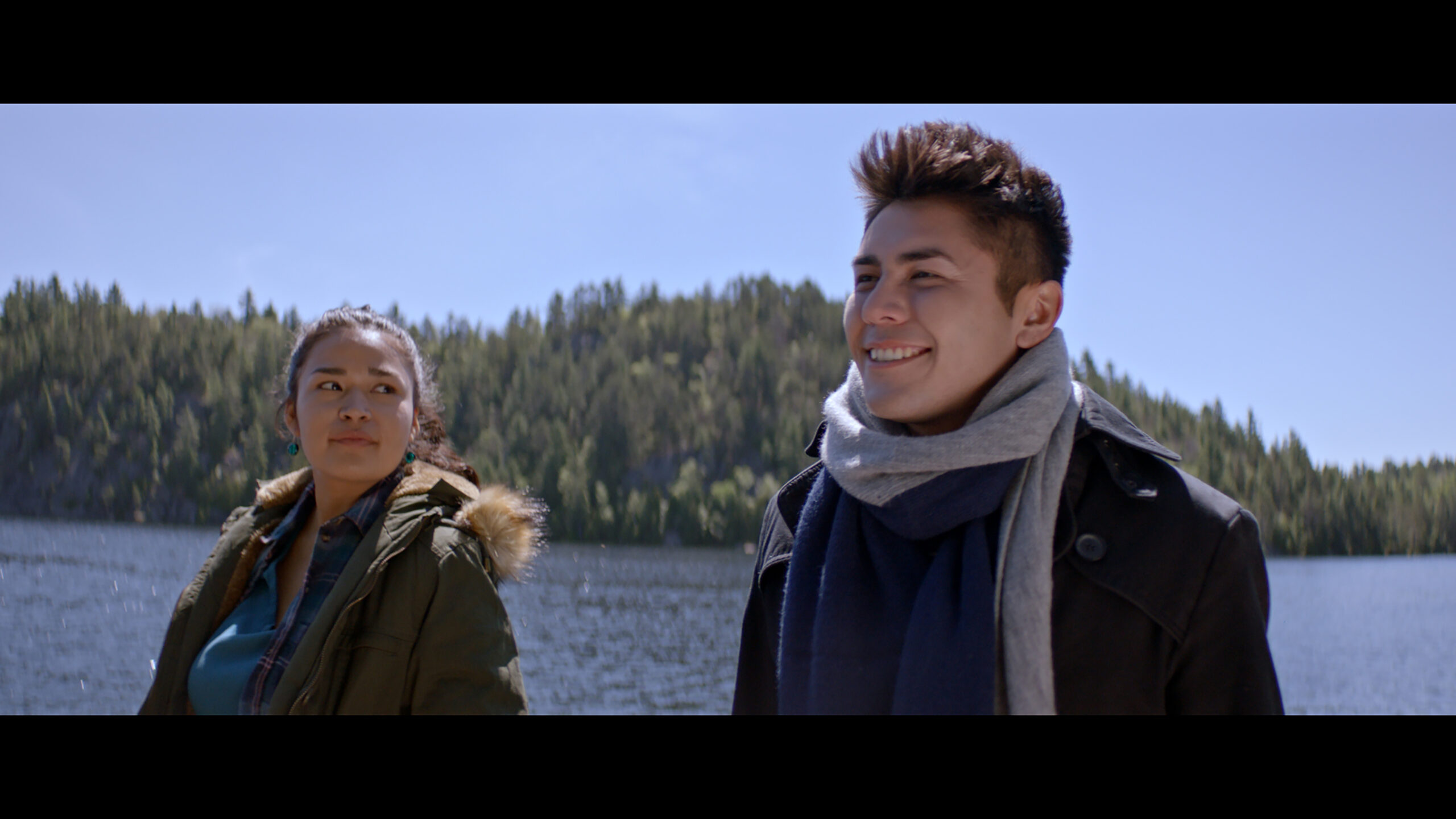 Two people are on a lake with a family of trees behind them. They are dressed for the chilly weather as they look at each other and smile.