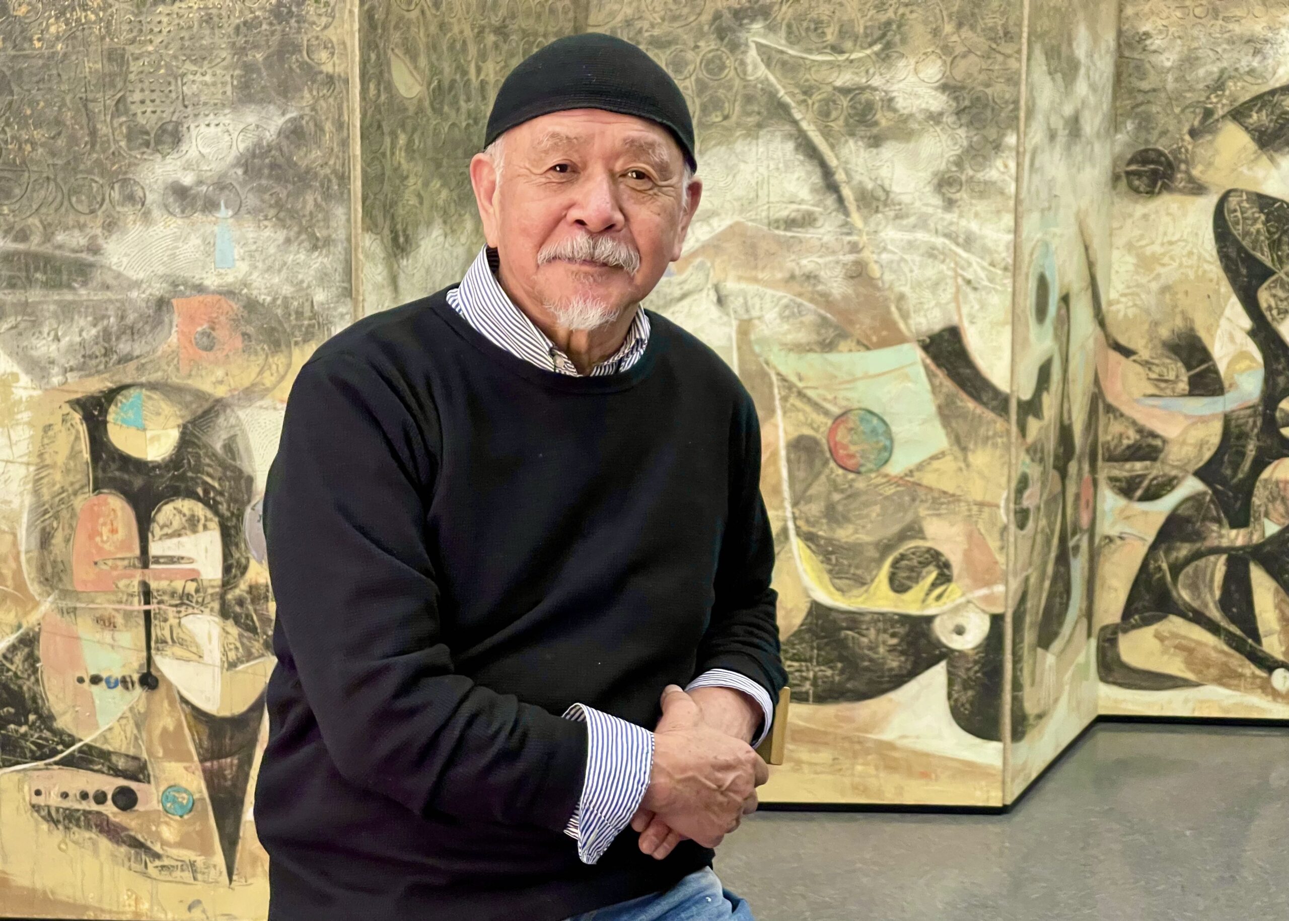 A person wearing a black cap and a black sweater looks to the camera to take a photo. Their artwork is displayed behind them.