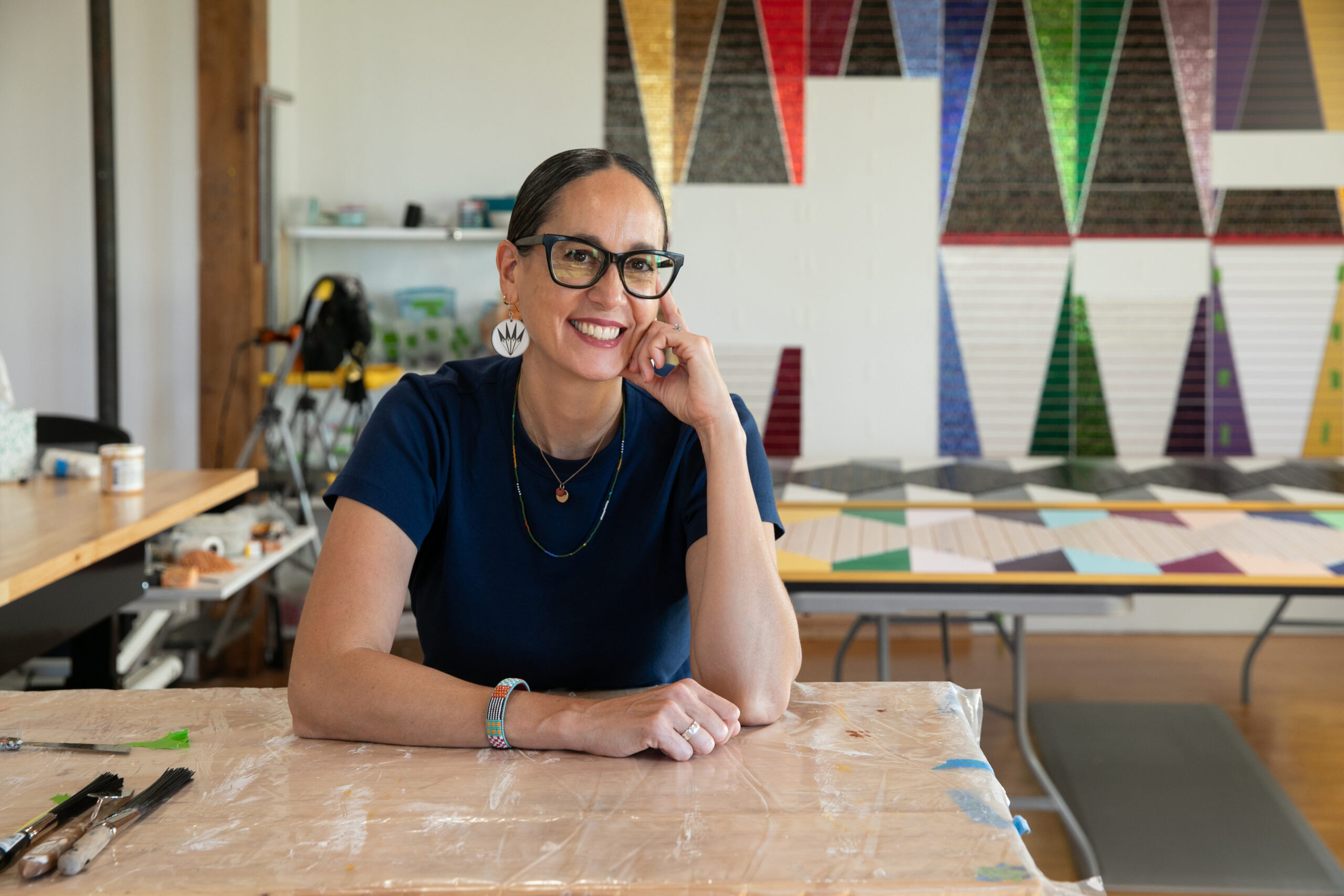 A person with black glasses and a navy blue shirt looks towards the camera to smile. They are wearing beaded Native jewelry.