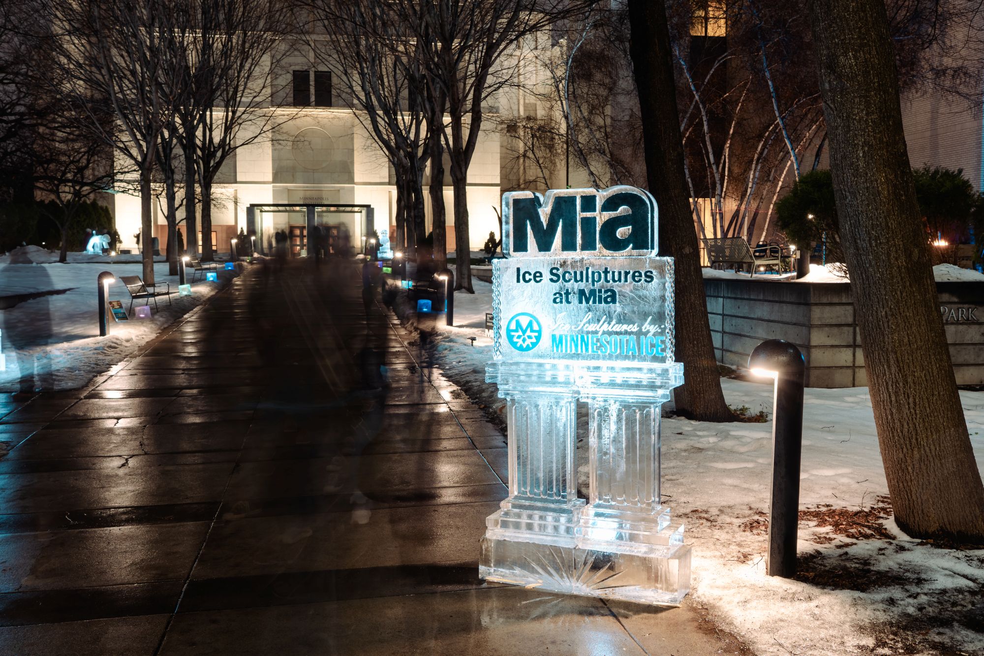 Ice sculpture sign for Mia at entrance pathway with snow and lights.