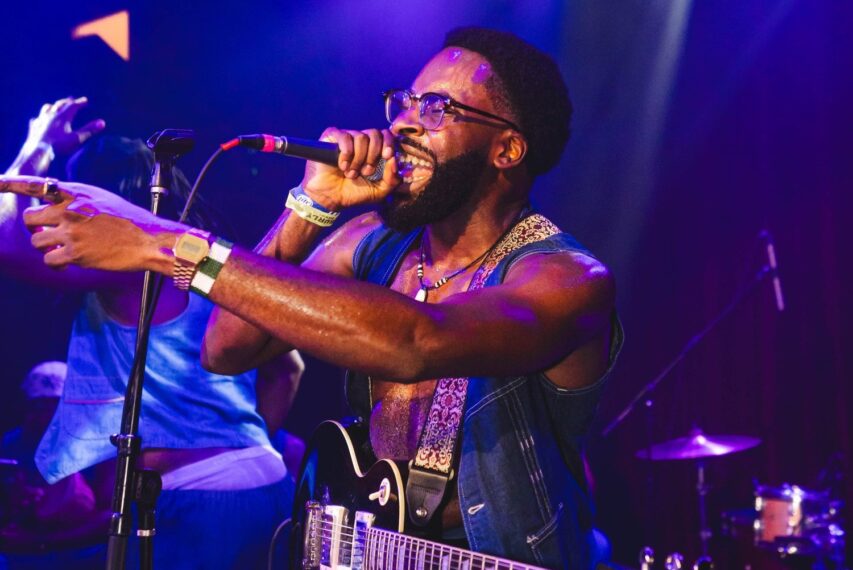 Musician singing on stage. He's holding a microphone in his right hand while wearing glasses and a sleeveless denim jacket. His guitar hangs from his neck and he appears to be passionately singing. 