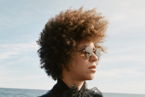 Person with curly hair wearing sunglasses against sky and sea background.
