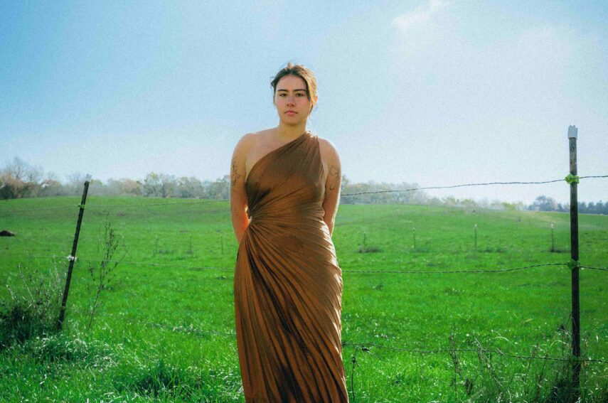 A person is standing in the center of a lush, green field. They are wearing a long, brown dress with a single shoulder strap. The background features a clear blue sky and the horizon is lined with distant trees. 