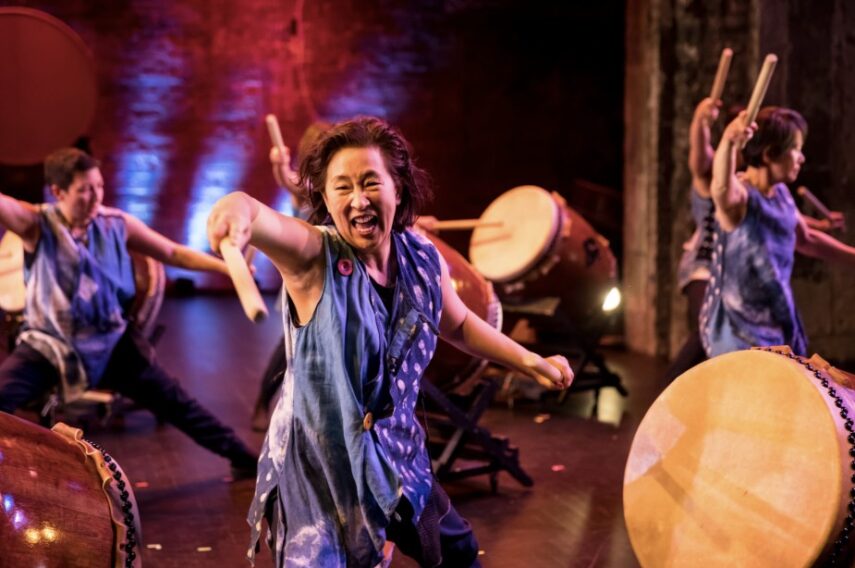 A group of people energetically performing on large traditional drums. They are on a dimly lit stage with colorful lighting, highlighting the drummers. The central figure is wearing a blue, sleeveless top and is striking a drum with wooden sticks, while appearing to shout or sing. Other drummers in the background are similarly dressed and fully engaged in the performance, each holding drumsticks in action.