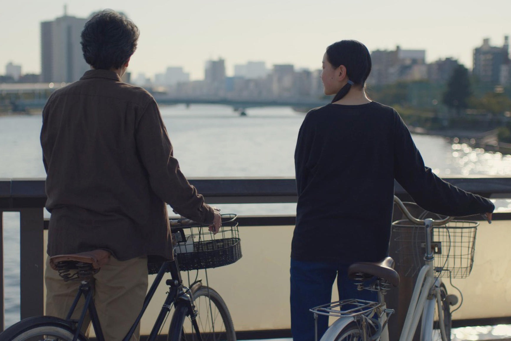 A young man and young women holding their bikes on a bridge overlooking a river in a city