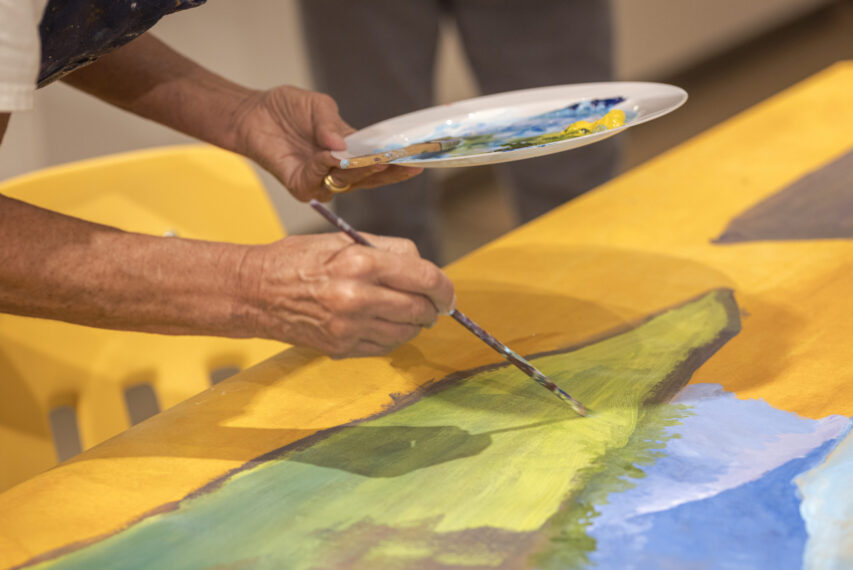 photo of an artist painting. One hand is holding a paint brush over a canvas of yellow, blue, and green hues, and the other hand is holding a plate with paints on it.