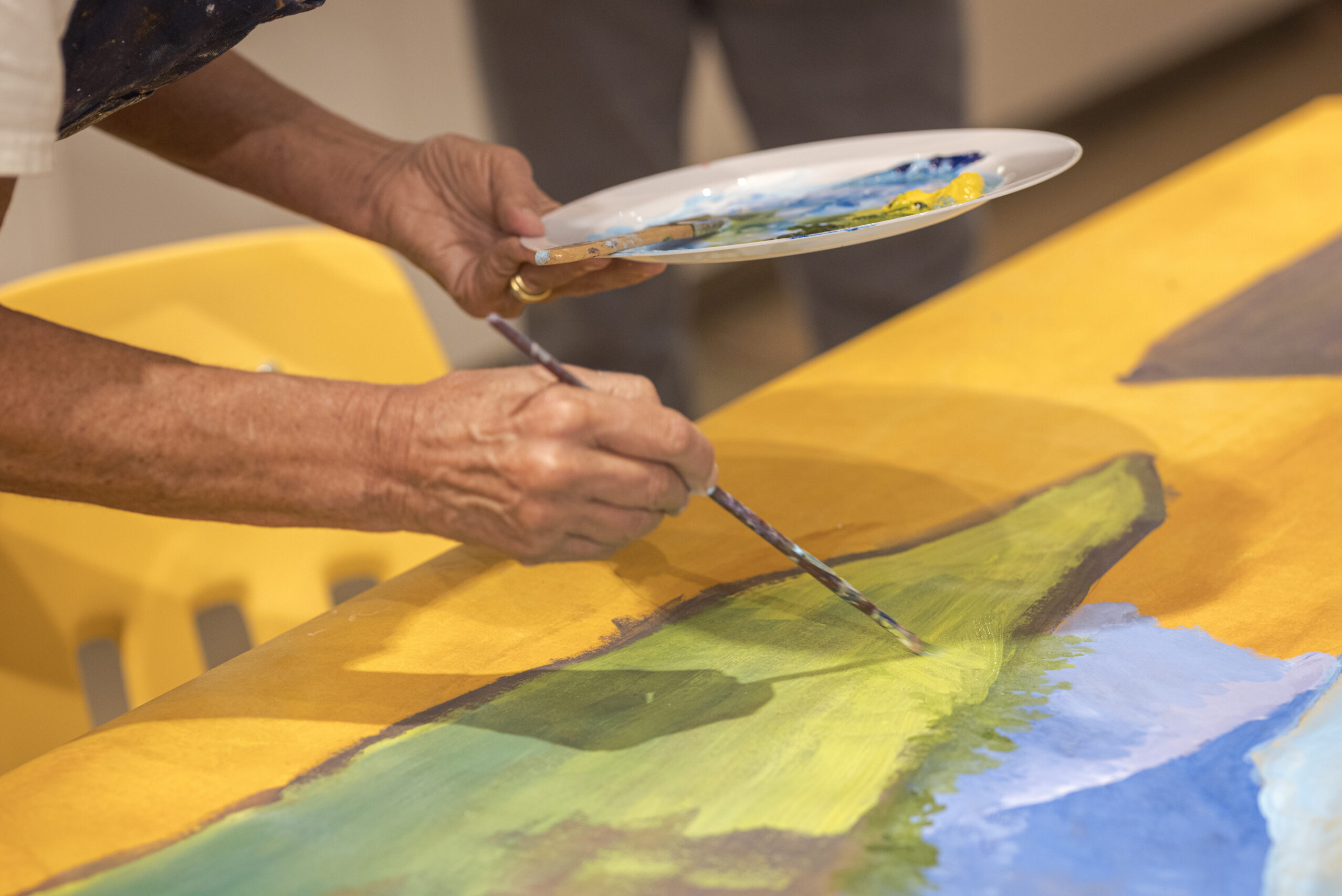 photo of an artist painting. One hand is holding a paint brush over a canvas of yellow, blue, and green hues, and the other hand is holding a plate with paints on it.