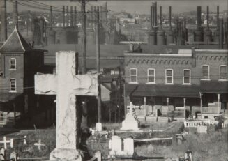 Black-and-white photograph of a cemetery with brick rowhouses and steel mills in the background