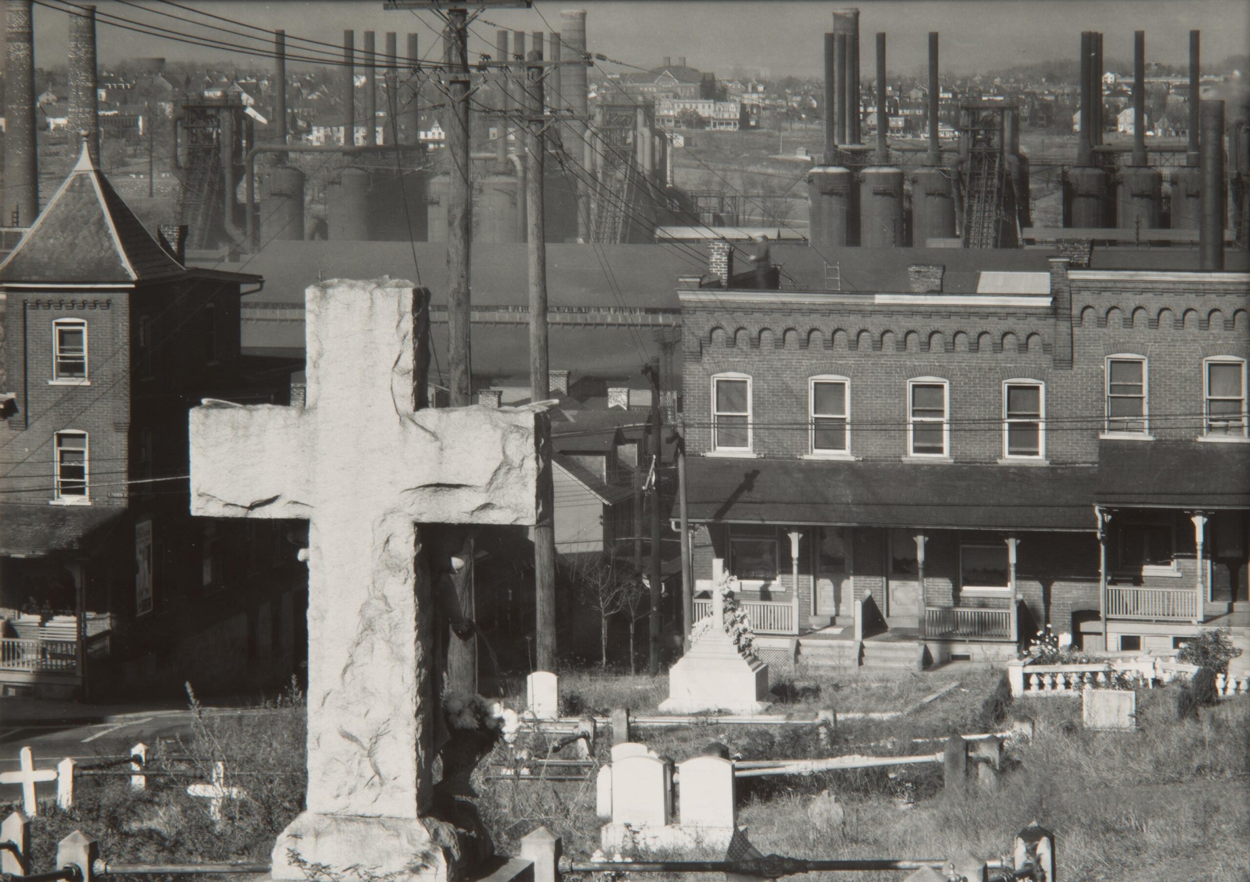 Black-and-white photograph of a cemetery with brick rowhouses and steel mills in the background
