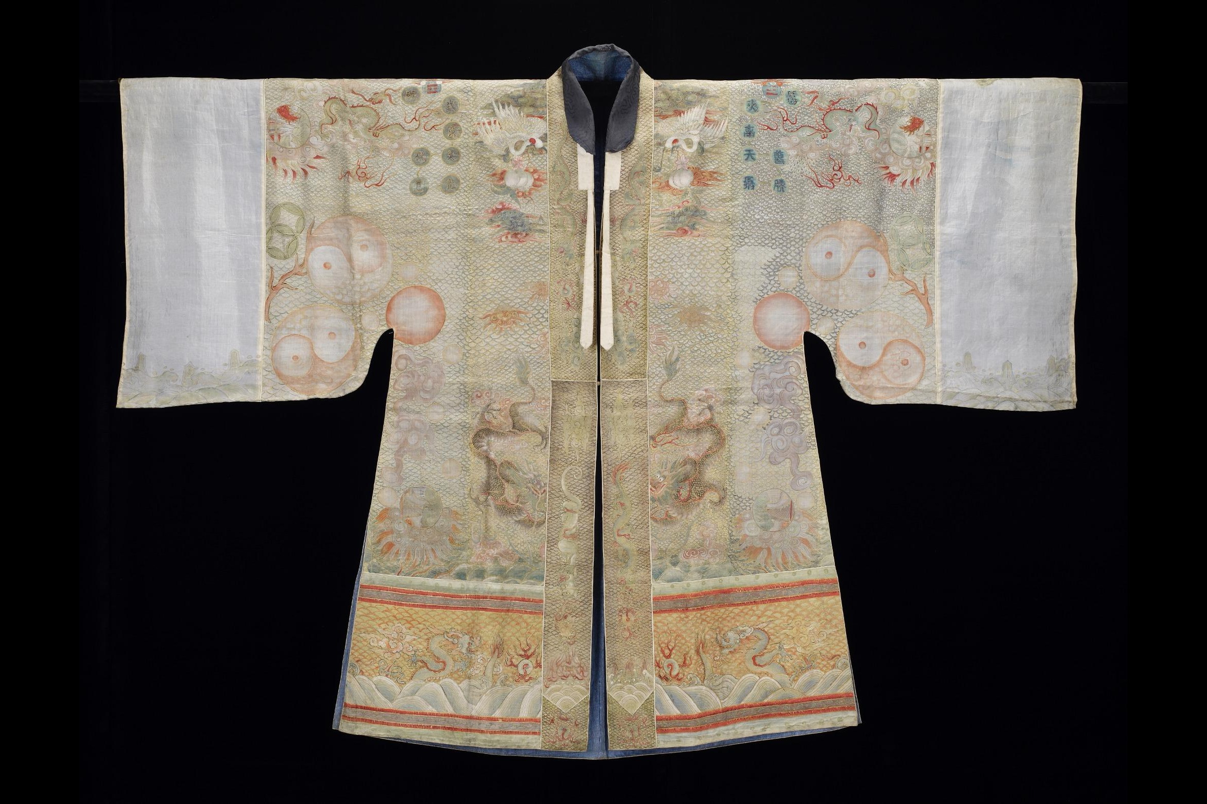 Photograph of an outstretch Daoist robe with long sleeves, with white fabric at the cuffs and beige fabric with orange, yellow, and brown embroidery on the body