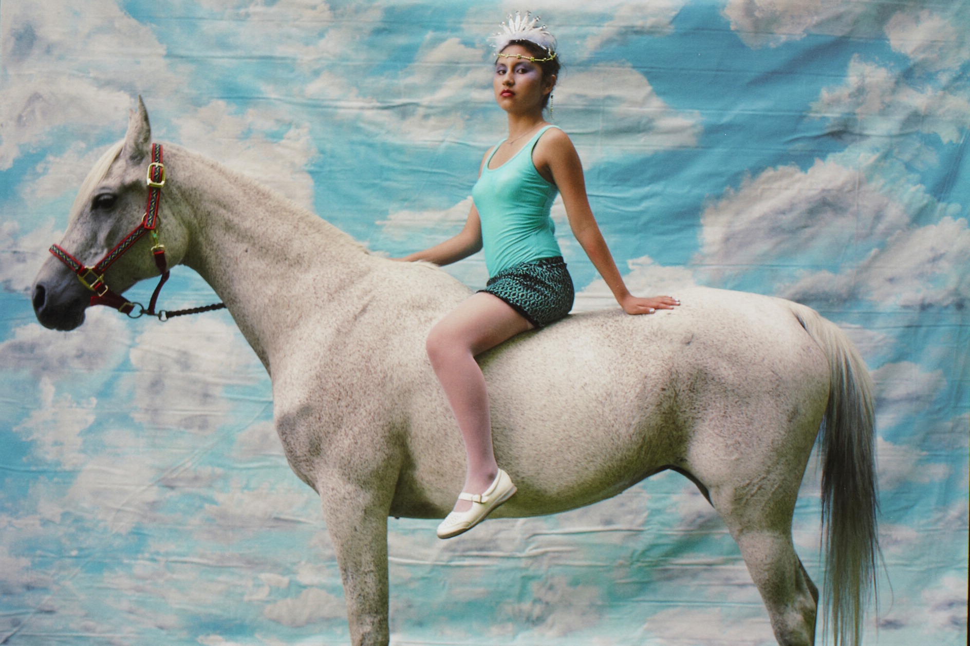 young woman wearing a turquoise tank top, turquoise and black skirt, white shoes and white beaded headdress, seated bareback on a white speckled horse, standing in front of a backdrop painted with blue sky and white clouds