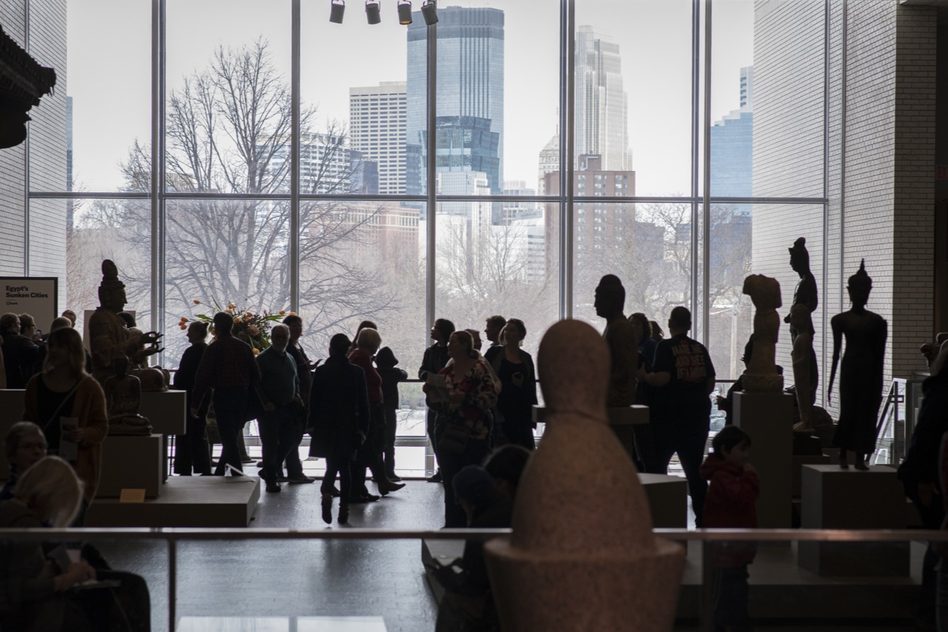 People walking around a Mia gallery, with a large glass window showing downtown Minneapolis in the background