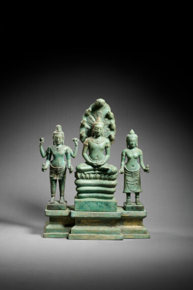 Bronze statue in a green hue of three gods; left: god with four arms standing; center: god seated cross-legged with a serpent headdress; right: god with two arms wearing a skirt standing 