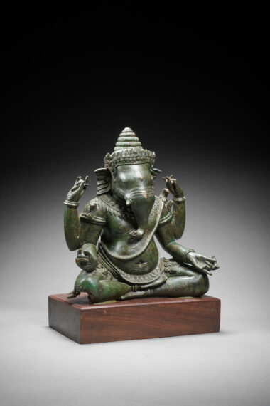 Bronze statue with a green hue of a god with an elephant head and four arms sitting cross-legged on the ground