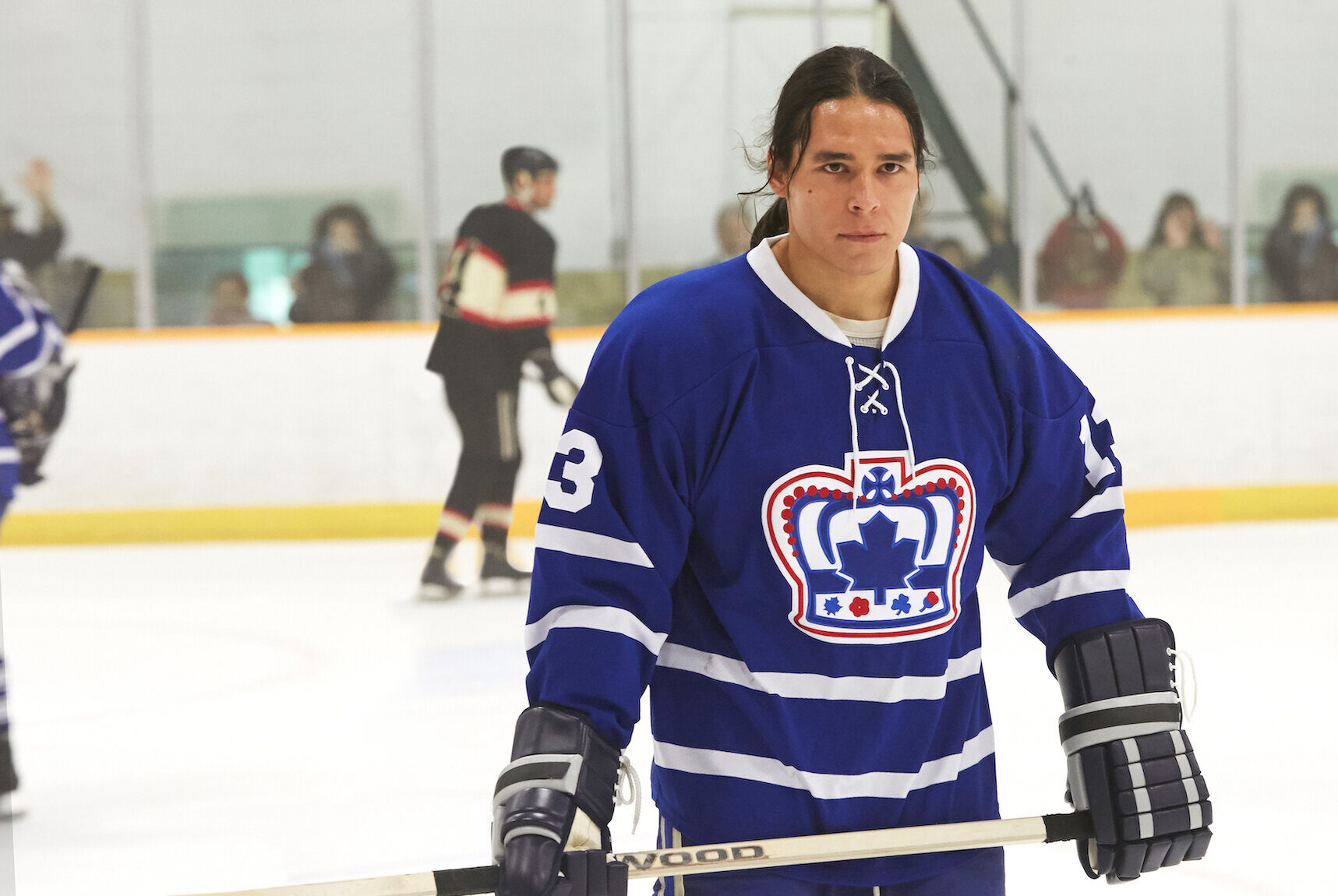 Photo of a young Native man wearing a blue hockey uniform and holding a hockey stick in an ice rink