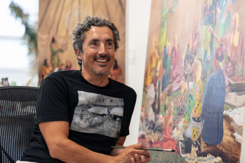 Photo of Kent Monkman sitting next to a painting in his studio holding a paint brush