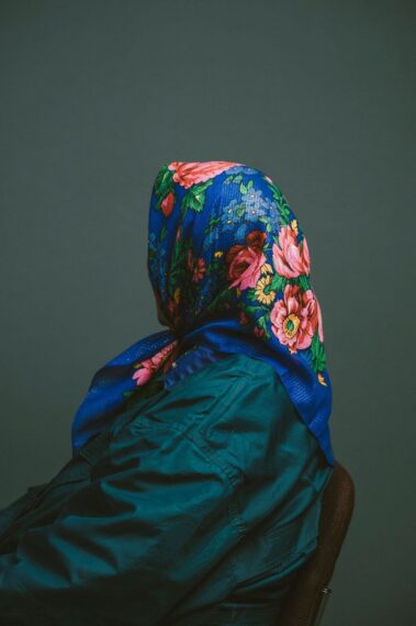 Photo of the profile a woman sitting, wearing a teal coat and a floral scarf wrapped around her head that obscures her face