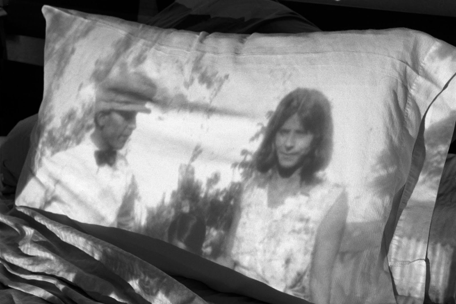 Black-and-white photograph showing an image of a man and a woman projected onto a pillow resting on a rumpled bed.