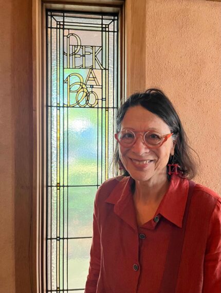 photo of a woman wearing a red shirt and red glasses standing in front of a stained-glass window that says "Peek a Boo"