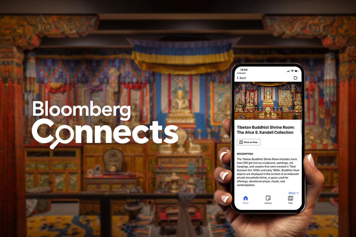 Hand holding a smartphone in front of Mia's Tibetan Buddhist Shrine Room. The smartphone screen displays information about the shrine room. The words 'Bloomberg Connects' are superimposed on the left side of the image.