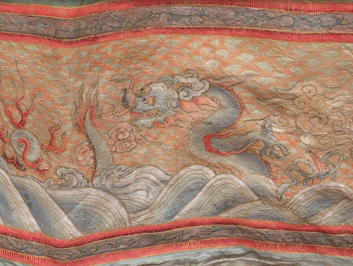 Ornate piece of fabric featuring an elaborate dragon design surrounded by swirling clouds and waves. The dragon has a long, serpentine body with distinct red accents. The fabric's colors include shades of orange, blue, and gray. 