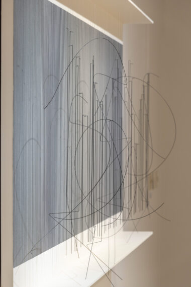 3D kinetic wall sculpture with dizzying optical effect; thin black and white vertical lines painted on background; in front are thin black wires hanging from nylon filament from white horizontal board across top edge; most wires are straight and vertical, but some are curved or sharply angled