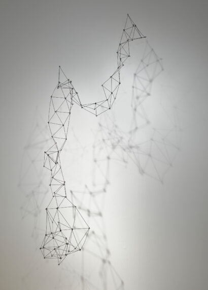 A wireframe sculpture hangs against a plain gray wall, forming abstract, geometric shapes. 
