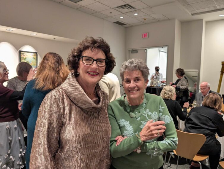 photo of two older women standing next to each with other people in the background