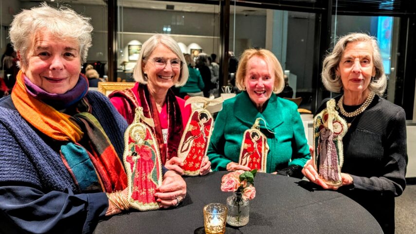 Photo of four older women standing next to each around a high-top table with a black tablecloth holding needle point angels