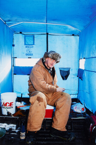 A man sitting inside a blue ice-fishing tent wearing a brown fur hat and brown overalls and holding a fishing rod.