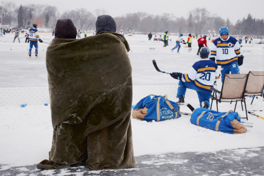 Outdoor ice rink with snow-covered surroundings. Two people stand on the rink wrapped in a large, brown blanket, facing away from the camera. They are watching a group of hockey players in blue, white, and gold uniforms.