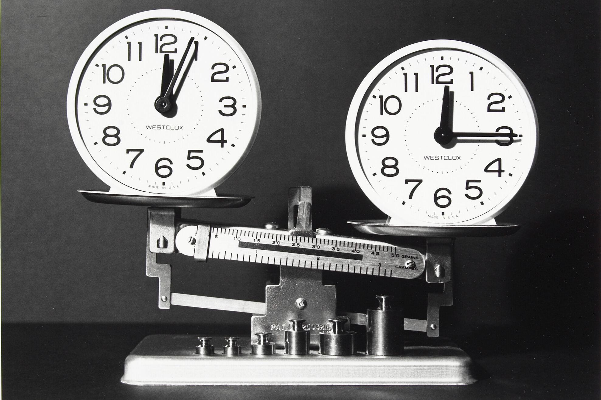 Two identical round clocks placed on opposite trays of a metal balance scale. Both clocks have white faces with black numbers and hands. The clock on the left shows the time at 12:04, while the clock on the right displays 12:15.