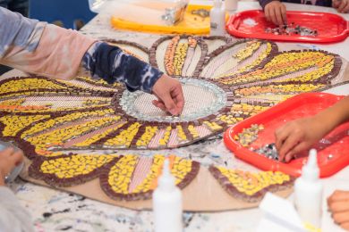 Children's hands working on a large, colorful mosaic project laid out on a table. The mosaic features a design resembling flower petals made from small pieces of yellow, orange, and brown materials.