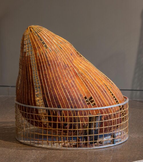 A large art installation resembling a woven basket. The structure has a rounded, organic shape with a mesh-like exterior, constructed from vertical and horizontal strips of a warm, earthy material in orange and brown hues. The top of the structure tapers to a point, similar to a large teardrop or gourd.