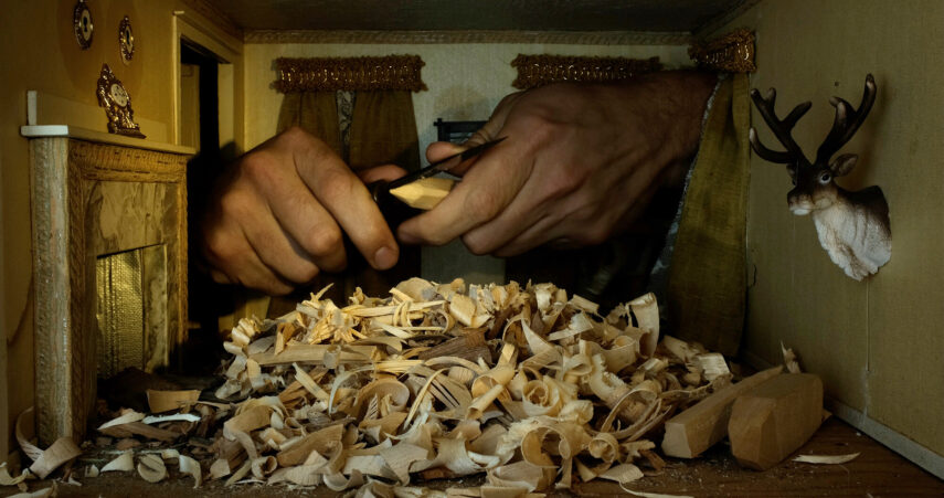 The image shows a pair of hands intricately carving a piece of wood. In the foreground, there's a pile of wood shavings scattered across the surface. The setting appears to be a small room or a miniature scene with a decorative wall featuring two framed ornaments and a small clock on a shelf. To the right, a mounted deer head decorates the wall. Curtains hang on the back wall, adding to the decor.