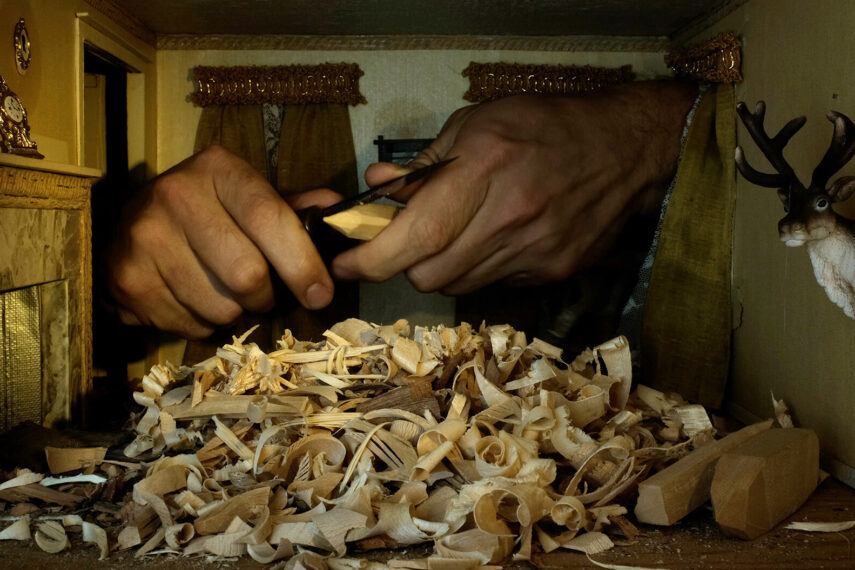 The image shows a pair of hands intricately carving a piece of wood. In the foreground, there's a pile of wood shavings scattered across the surface. The setting appears to be a small room or a miniature scene with a decorative wall featuring two framed ornaments and a small clock on a shelf. To the right, a mounted deer head decorates the wall. Curtains hang on the back wall, adding to the decor.