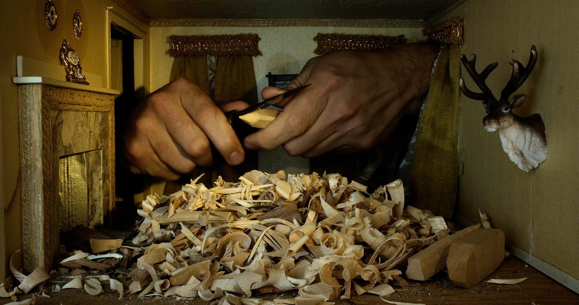 The image shows a pair of hands intricately carving a piece of wood. In the foreground, there's a pile of wood shavings scattered across the surface. The setting appears to be a small room or a miniature scene with a decorative wall featuring two framed ornaments and a small clock on a shelf. To the right, a mounted deer head decorates the wall. Curtains hang on the back wall, adding to the decor.