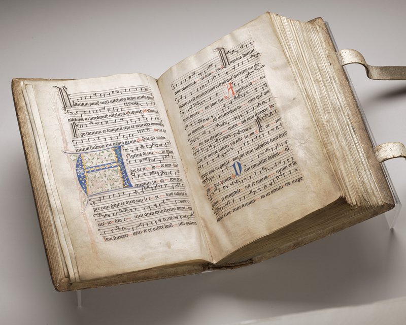 Open medieval illuminated manuscript showing two facing pages of large, hand-written Latin musical notation and text in black and red ink on vellum, with decorative initials and ornate embellishments in red and other colors, bound in a thick, aged leather cover with visible wear and clasps.