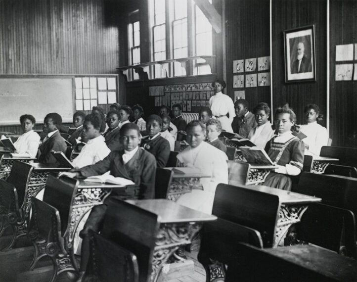 Classroom with students seated at desks and a teacher standing by the wall.