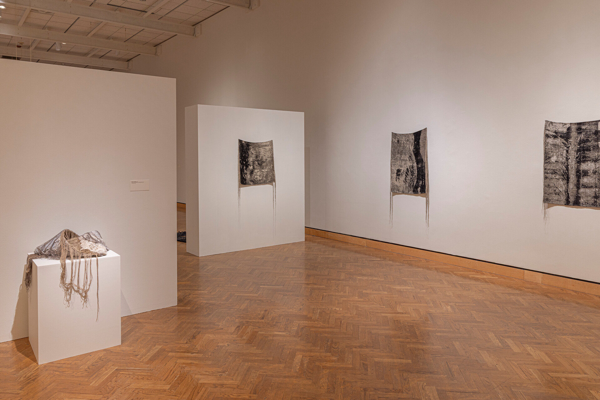 View of a gallery with woven textiles on walls and a pedestal.