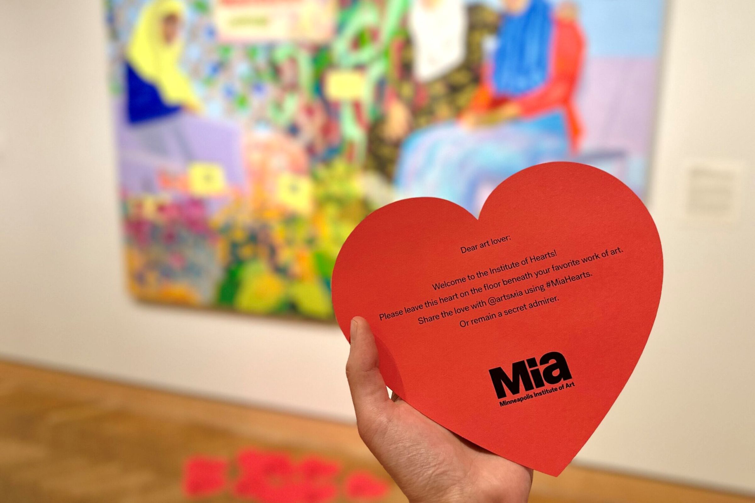 Photo of someone holding a red, heart-shaped piece of paper in front of a work of art. Other red hearts are on the floor in front of the artwork. These hearts are used during the Institute of Hearts event during the Valentine’s season.