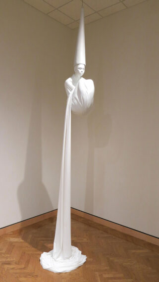 Sculpture of a figure with a tall, pointed hat suspended from the ceiling. The figure appears to be wrapped in flowing white fabric that gently drapes and pools on the wooden floor. 